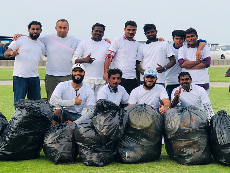 A group of volunteers in casual attire posing with collected trash bags during a community cleanup event outdoors, smiling and giving thumbs up.