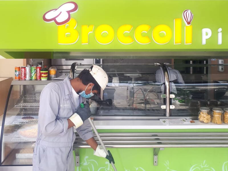A worker in a grey uniform and white cap cleaning the front area of a green food stall labeled 'Broccoli' with cans of soda and jars visible on the counter. The worker is wearing gloves and a face mask while sweeping.