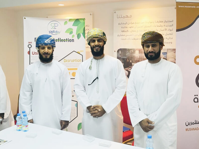Three men in traditional Omani attire standing together at an exhibition booth with informational banners in the background, smiling for the photo