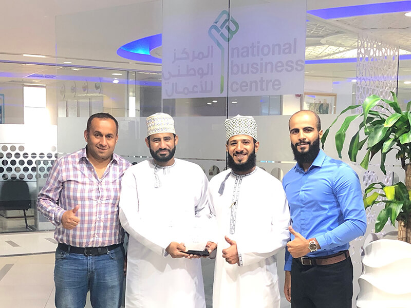 Four men standing together at the National Business Centre, with two individuals in traditional Omani attire holding an award, while the others are dressed in casual and business attire, smiling and giving thumbs up.