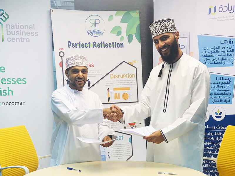Two men wearing traditional Omani clothing shaking hands in front of Perfect Reflection banners promoting a business partnership, holding documents, and smiling during a signing event.