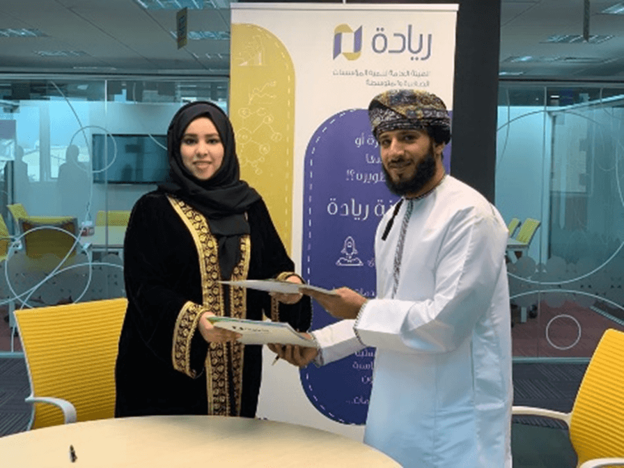 Two professionals exchanging documents in a formal setting, a woman in a traditional black abaya and gold-trimmed scarf on the left, and a man in an Omani dishdasha and turban on the right. A banner behind them displays Arabic text and corporate logos.